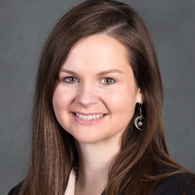 Keynote speaker Kacie St. George smiling in a professional headshot with long brown hair, wearing a black blazer and silver crescent moon earrings