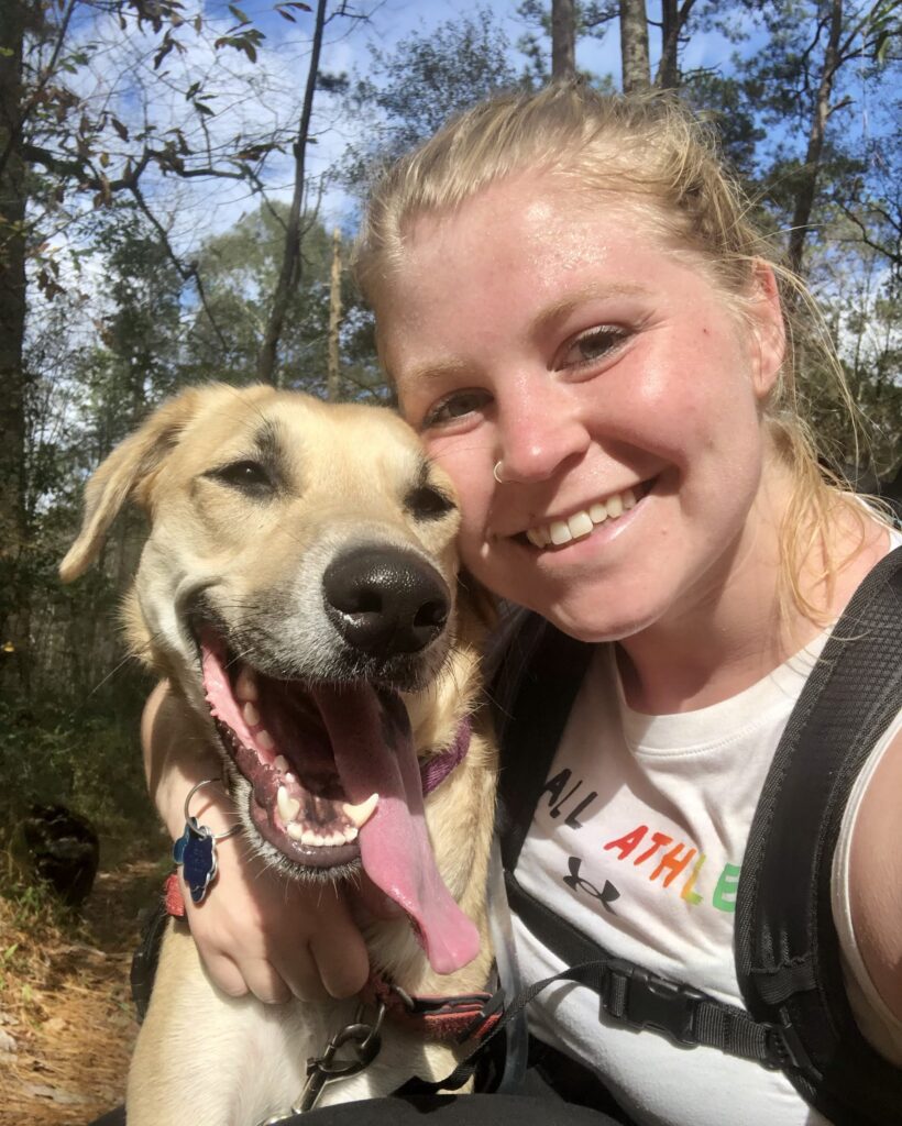 Tess is in a forest with her dog, a blond lab-mix, and both are smiling