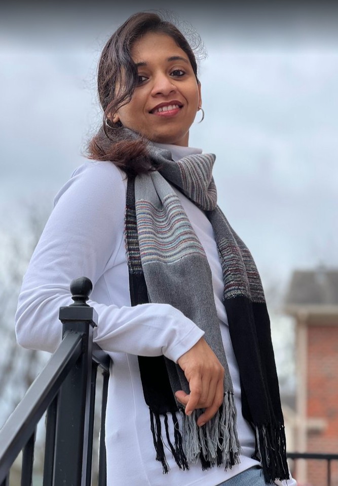 Mira in long sleeve shirt and scarf, outside on a cloudy day leaning against a railing