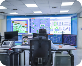 A person sitting at a multi-monitor workstation in a control room, facing a large wall-mounted display showing various system dashboards, maps, and operational data screens in a blue-lit industrial or infrastructure monitoring facility.