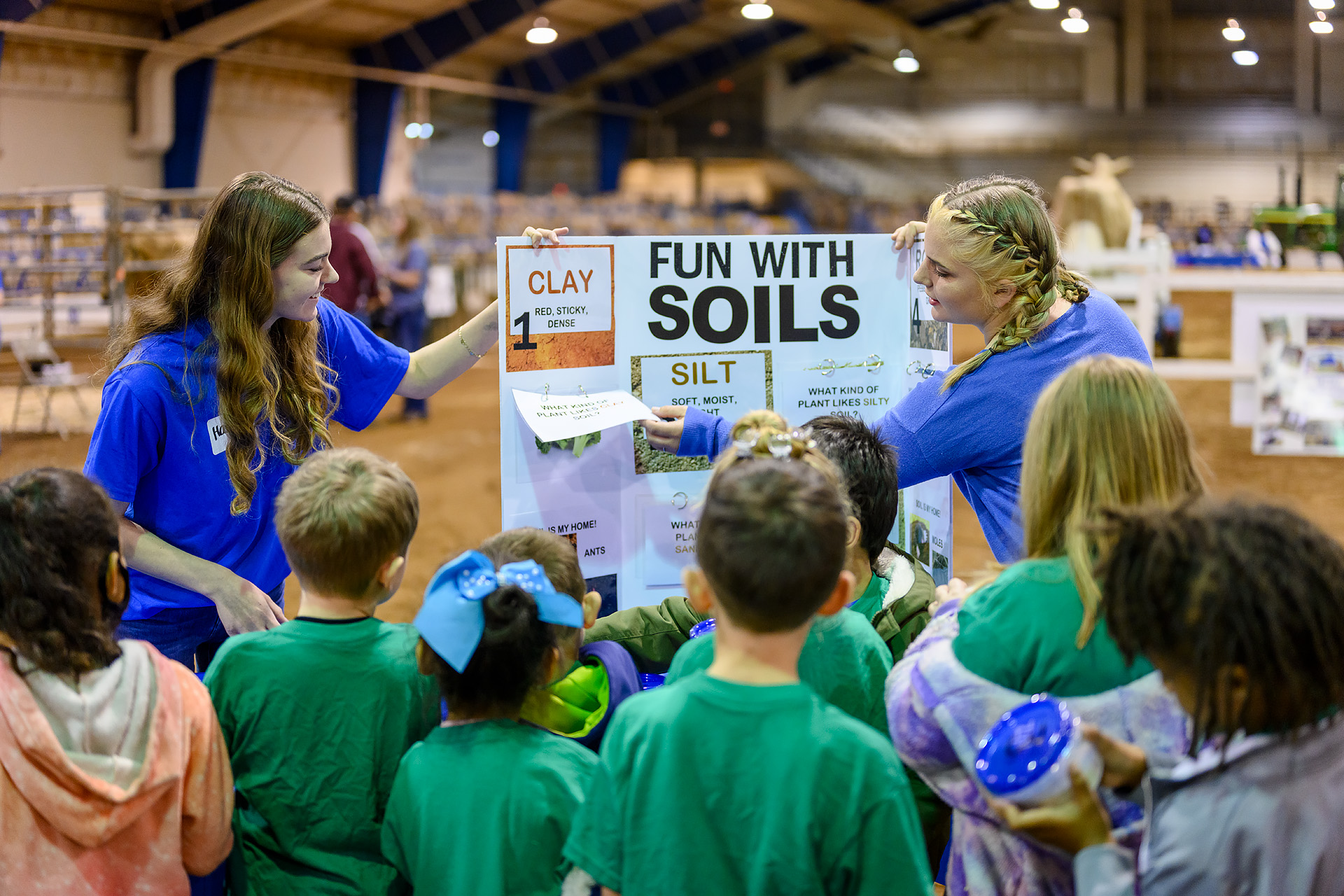 Two young women in blue shirts hold up a 'Fun with Soils' educational poster about clay, silt, and other soil types while a group of young children gathered around them look on attentively, inside a large indoor arena.