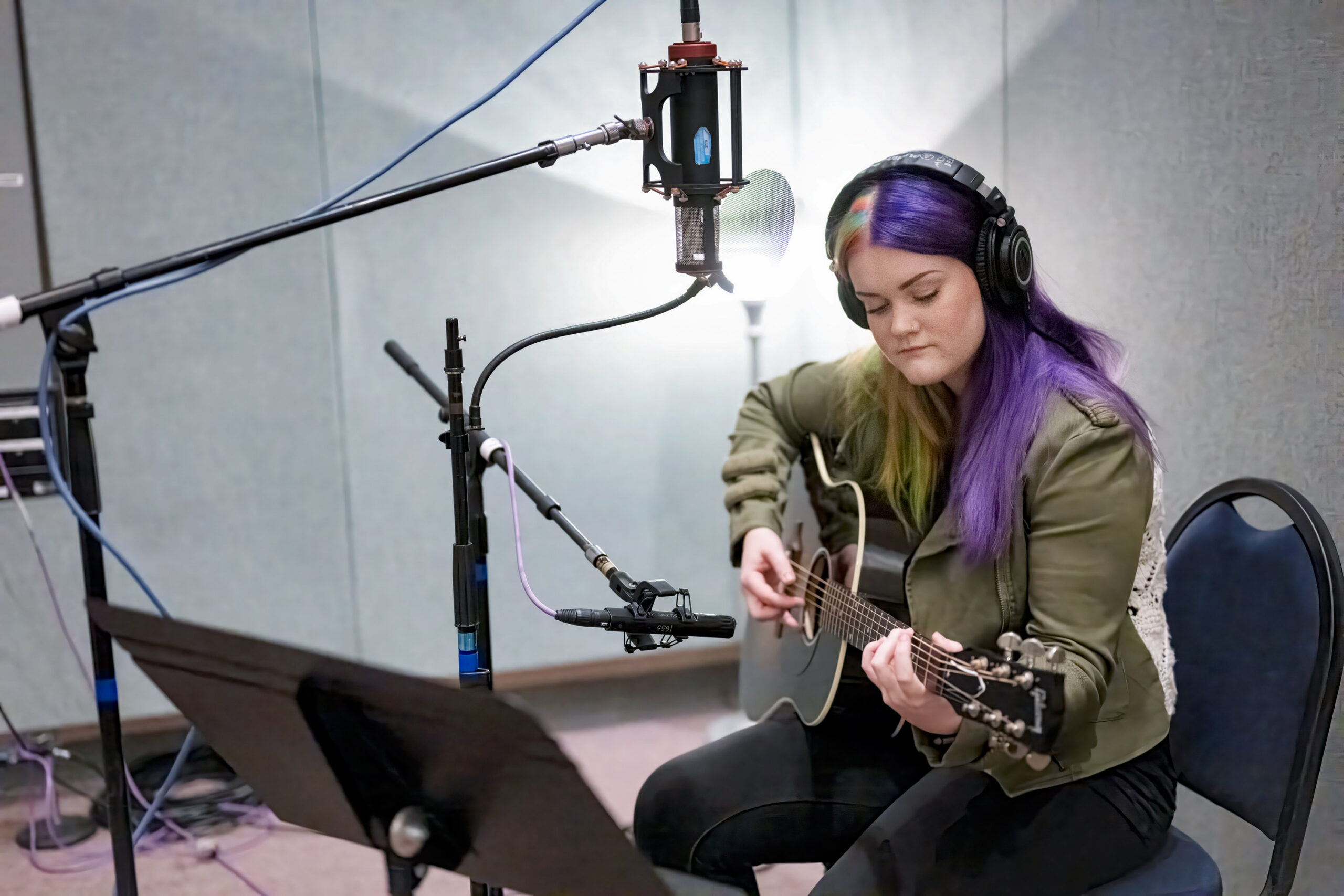 A woman with purple and green hair wearing headphones plays an acoustic guitar while seated in a professional recording studio, surrounded by condenser microphones and a music stand.