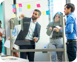 A group of business professionals collaborating in a modern office, writing and reviewing notes on a glass whiteboard covered with colorful sticky notes.