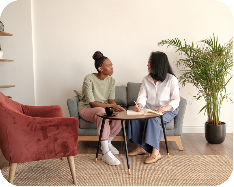 Two women sit together on a gray sofa in a warm, welcoming room, engaged in conversation while one takes notes in a notebook, suggesting a counseling or advising session.