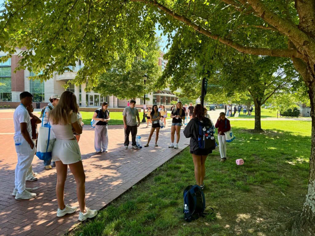 Students attending orientation standing in the shade of a tree.