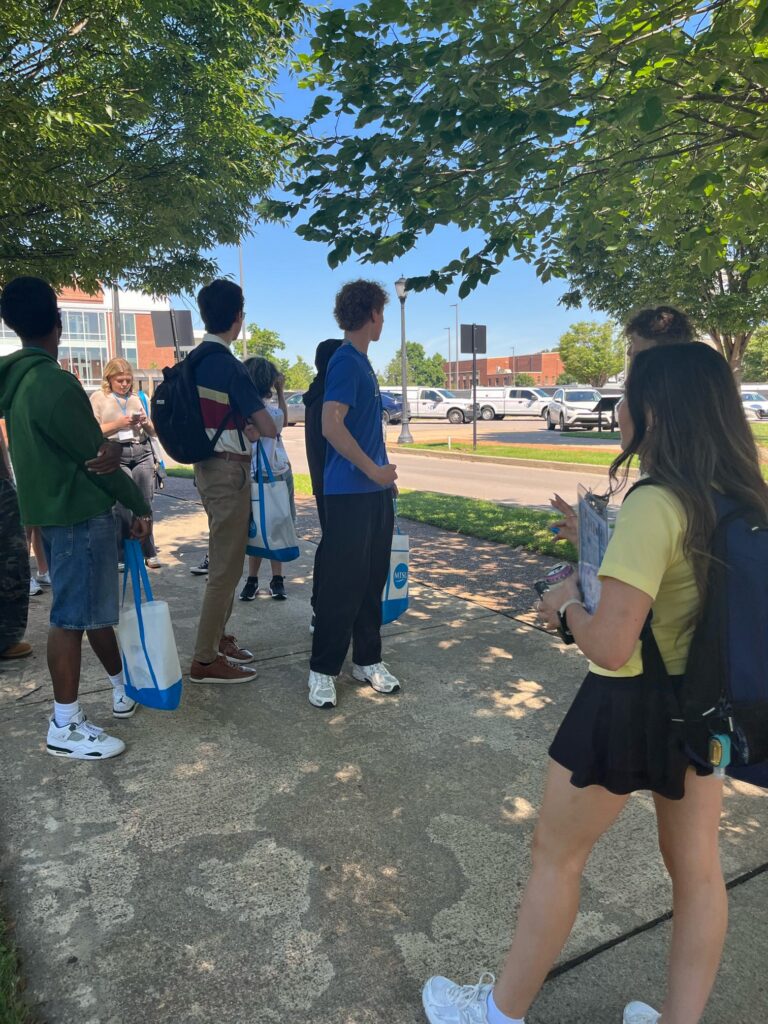 Students attending orientation standing in a shaded area while their Student Orientation Assistant leads a conversation.