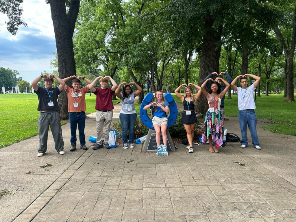Students attending orientation posing for a photo at the blue horseshoe forming the shape of a heart with their hands and arms with their Student Orientation Assistant.