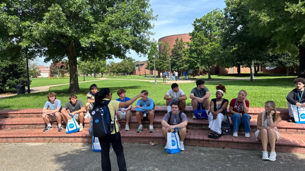 Students attending orientation sitting outside while their Student Orientation Assistant points to a building.